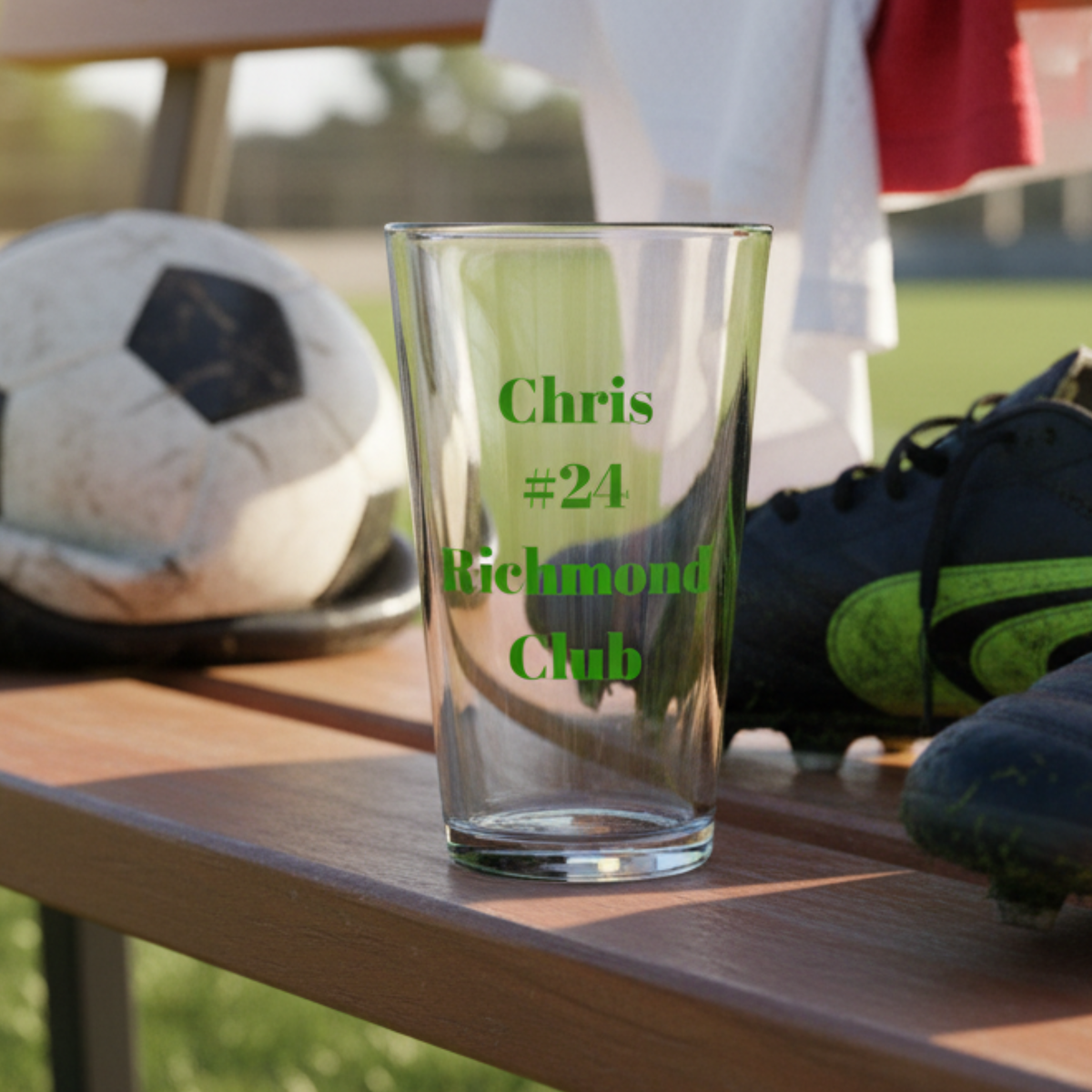 Pint glass engraved with sample personalization reading "Chris #24 Richmond Club" in green lettering, displayed on wooden table with soccer ball and cleats in background.