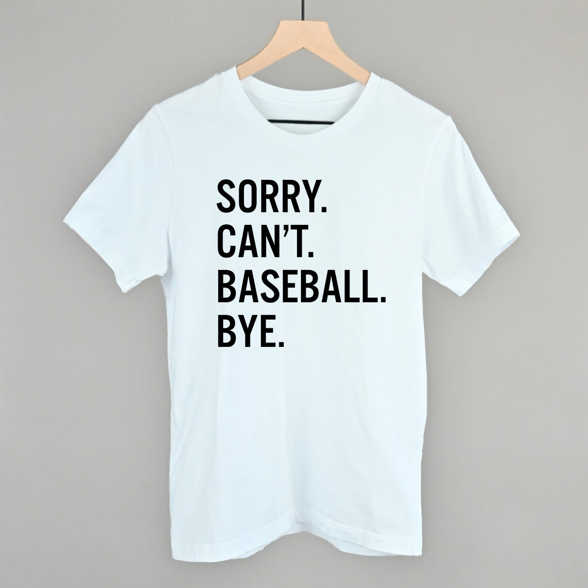 White short-sleeve t-shirt with black printed text reading "SORRY. CAN'T. BASEBALL. BYE." displayed on wooden hanger against gray background