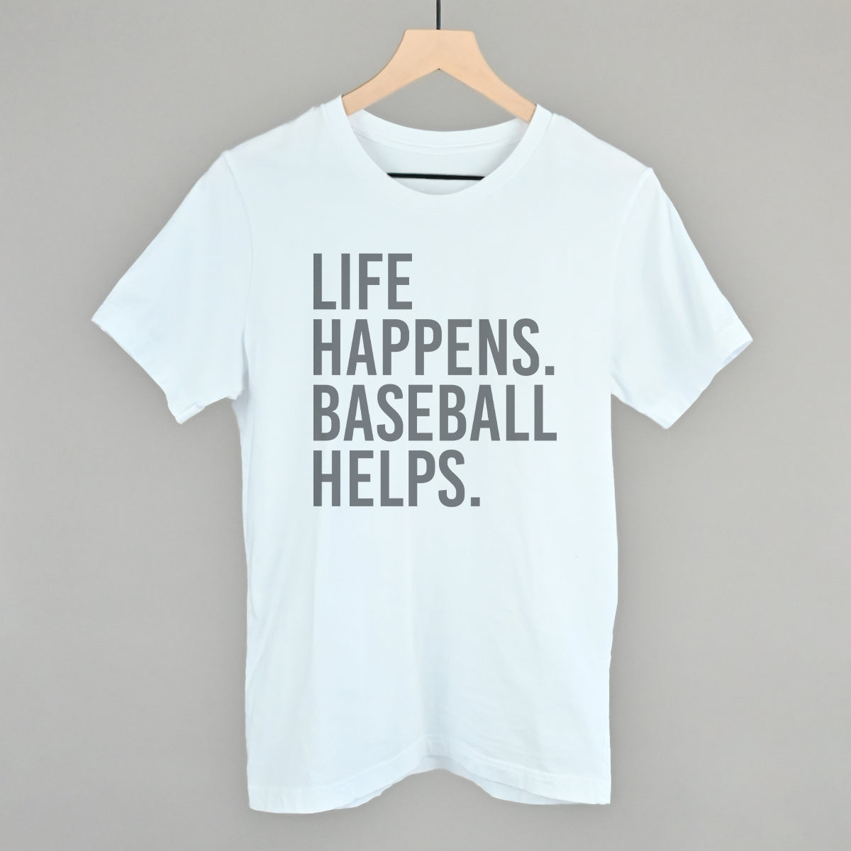 White t-shirt with gray printed text reading "LIFE HAPPENS. BASEBALL HELPS." displayed on wooden hanger against gray background.