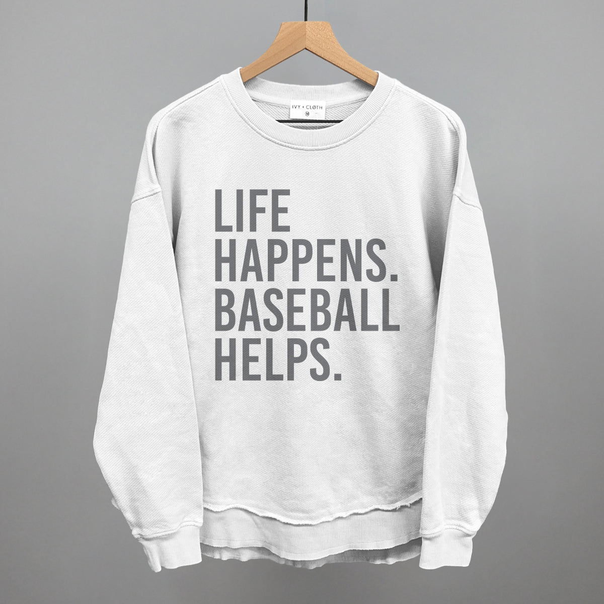 White crewneck sweatshirt with gray printed text reading "LIFE HAPPENS. BASEBALL HELPS." displayed on wooden hanger against gray background.