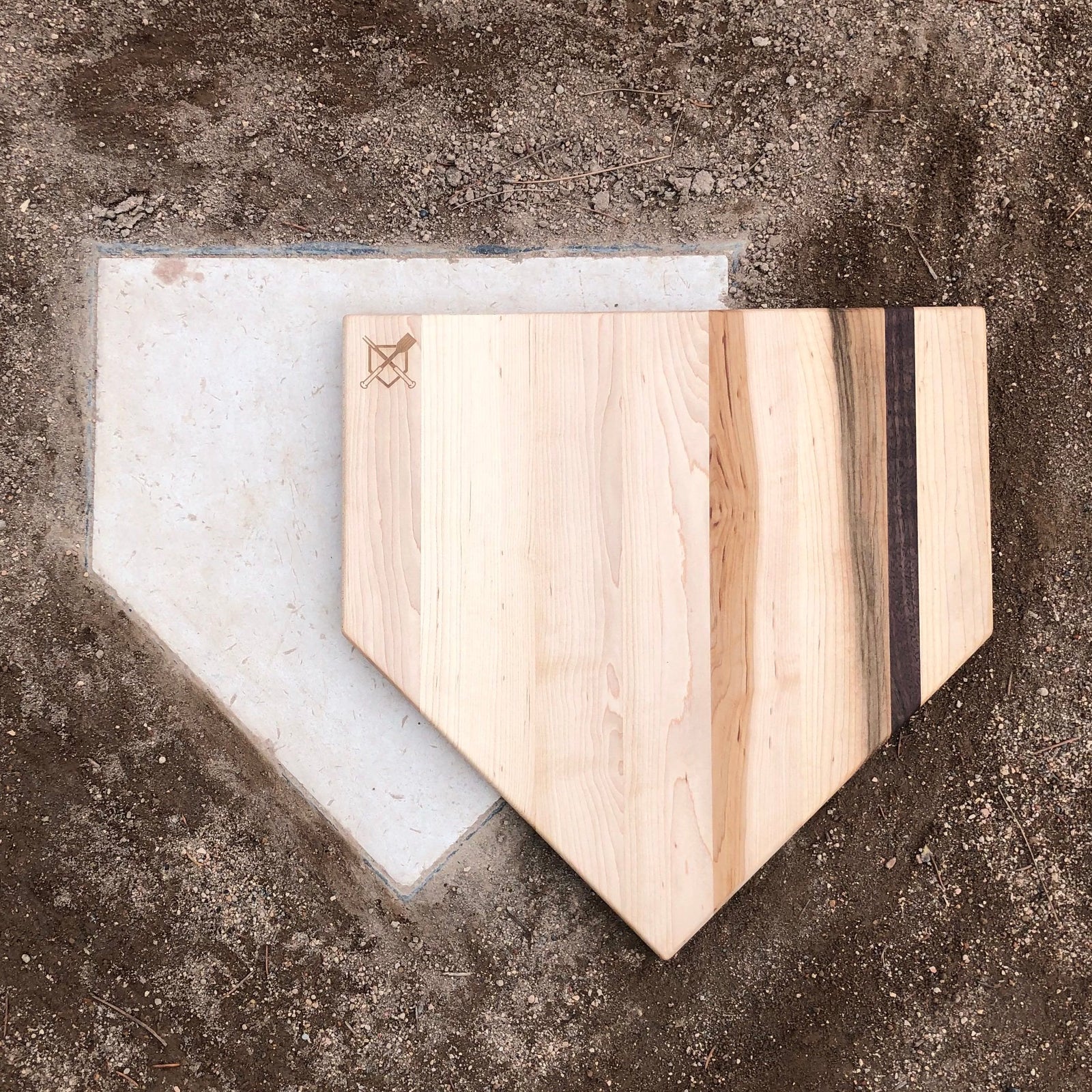Home plate-shaped cutting board in light wood with black walnut accent stripe, displayed on concrete next to white template showing size comparison