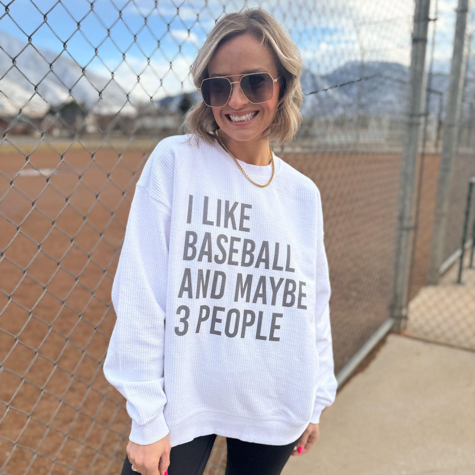 Woman wearing white crewneck sweatshirt with gray text reading "I LIKE BASEBALL AND MAYBE 3 PEOPLE," standing at a baseball field behind chain-link fence.