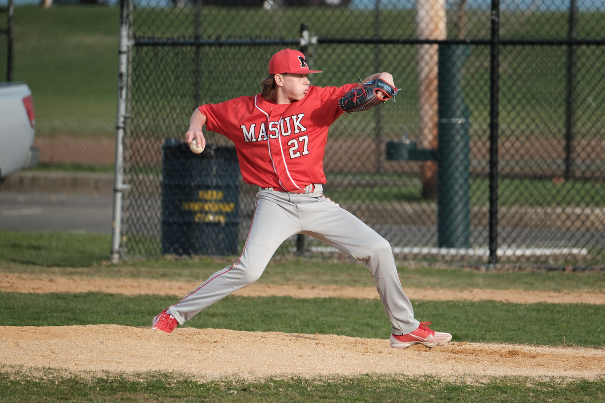 A high school pitcher in a red Masuk number 27 jersey mid delivery on the mound, the kind of travel ball weekend Batter Box Sports gear is picked for