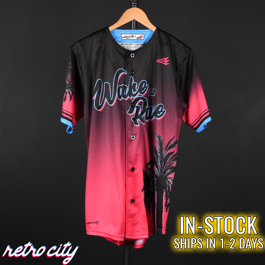 Black and hot pink gradient baseball jersey with "Wake and Rake" text, palm graphics, blue striped sleeves, and blue interior neckline, displayed on wooden hanger.