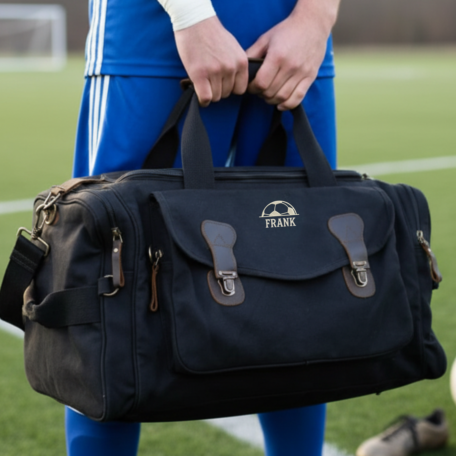Black canvas duffle bag with brown leather accents and brass hardware, personalized with sample name "FRANK" and soccer logo, held by person in blue athletic wear on soccer field