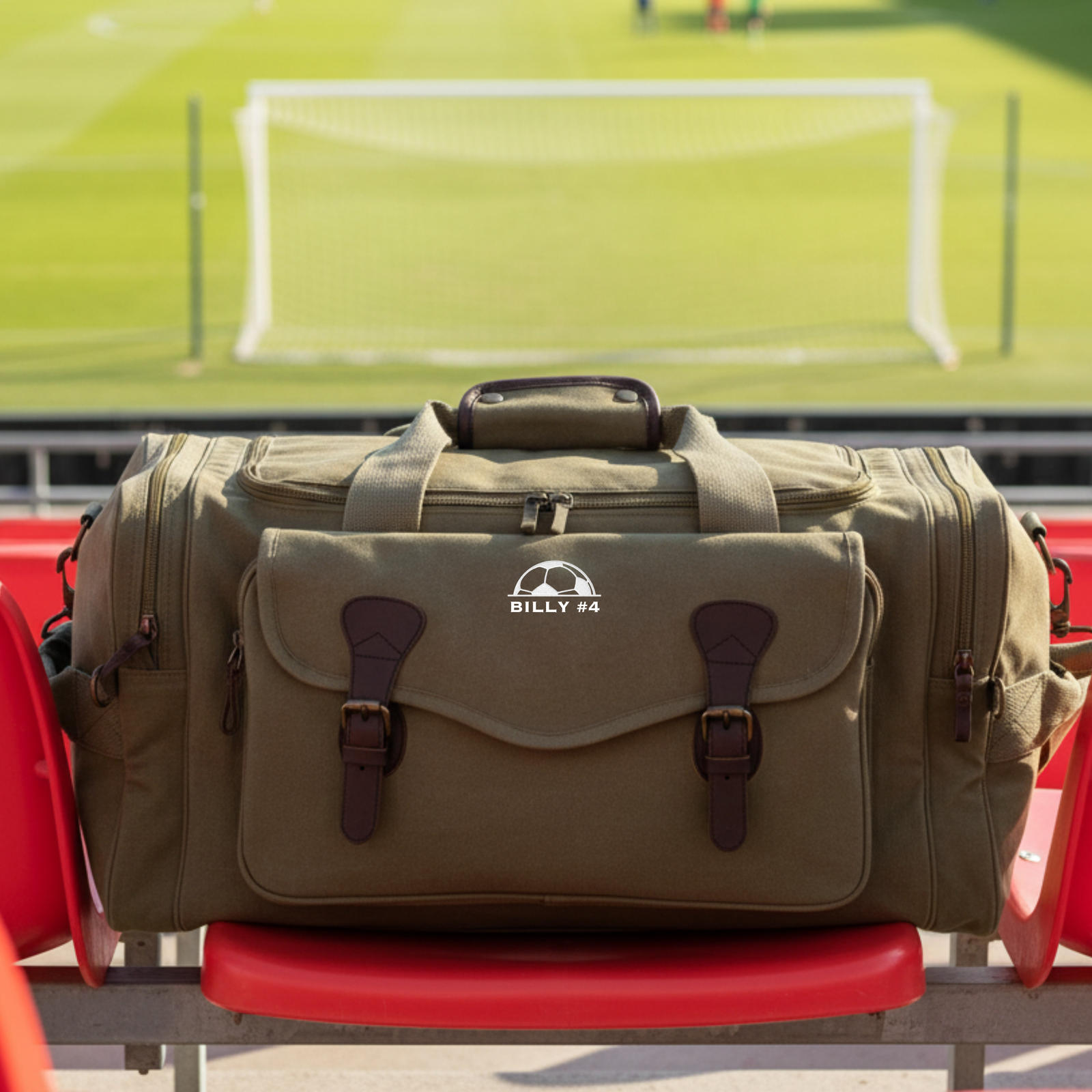 Tan canvas duffle bag with dark leather straps and brass hardware, personalized with sample name "BILLY #4" and soccer ball emblem, shown on red bench at soccer field