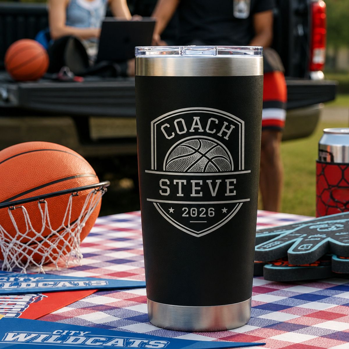 Basketball coach tumbler engraved with a Coach Steve 2026 basketball shield on a checkered table beside a basketball hoop at a team party outdoors