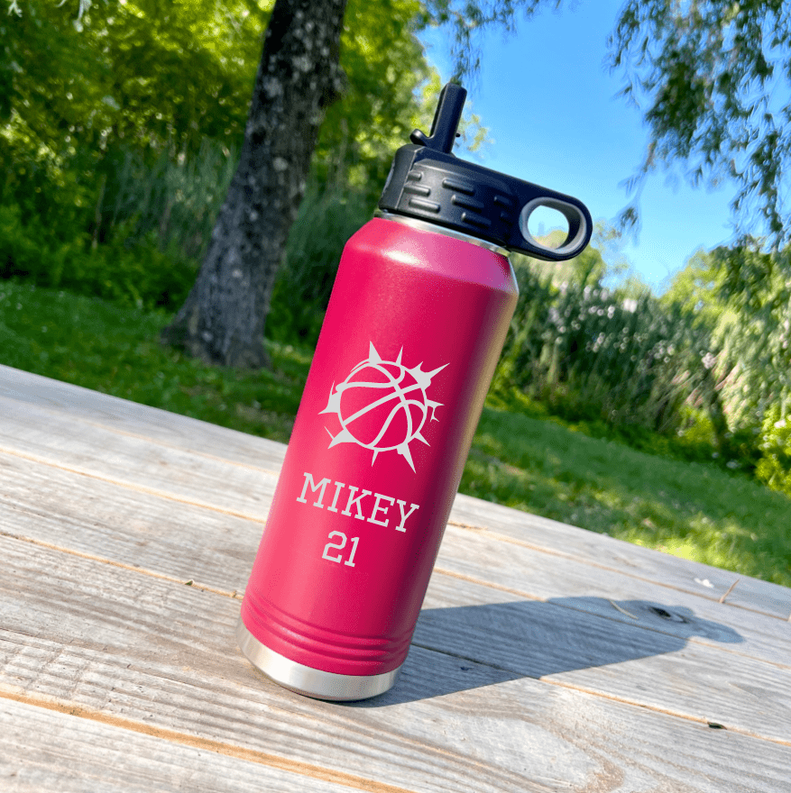 Pink personalized basketball water bottle with a black straw lid, laser engraved with a basketball, the name Mikey and the number 21, standing on a picnic table outside