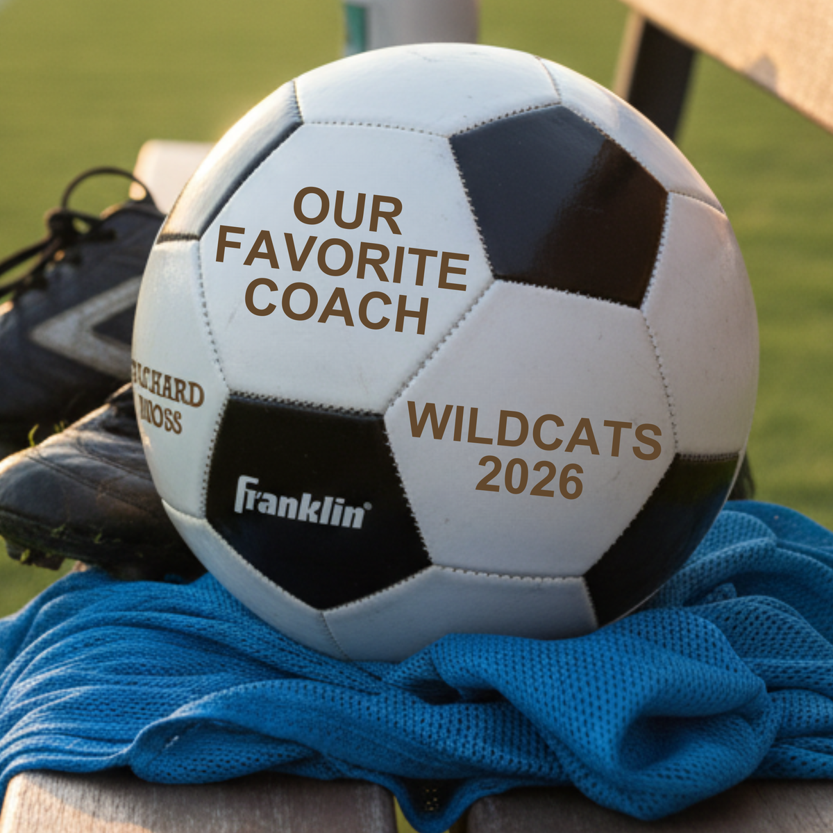 White and black soccer ball with laser-engraved sample personalization reading "OUR FAVORITE COACH," "RICHARD HROSS," and "WILDCATS 2026" in brown text, displayed on blue fabric with soccer cleats and grass field in background.