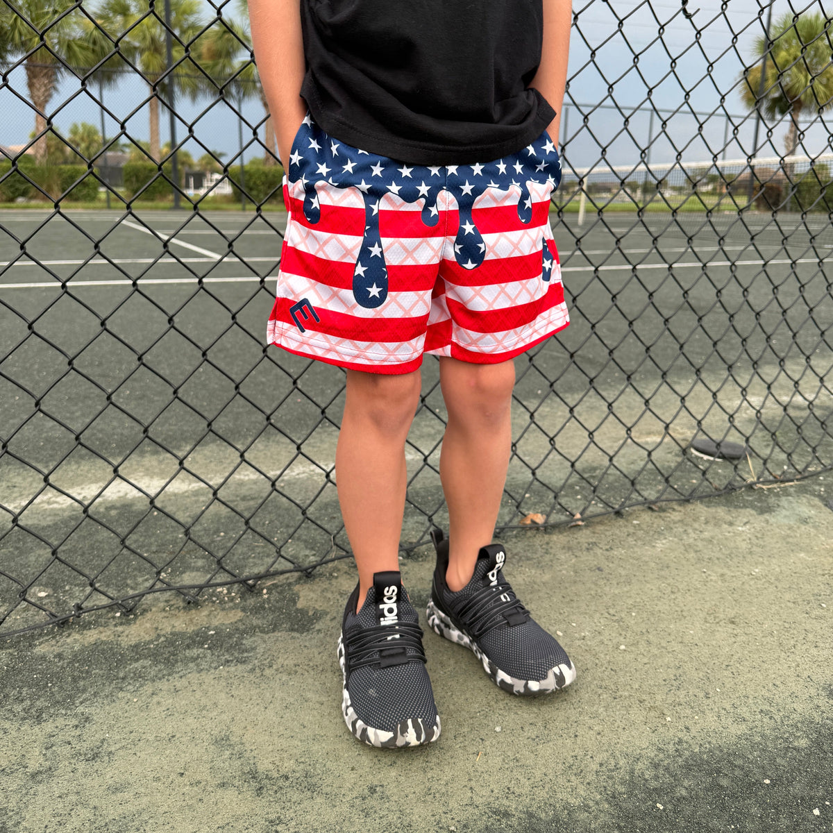 Child wearing red, white, and navy USA flag-patterned shorts with star and stripe design, standing at baseball field fence