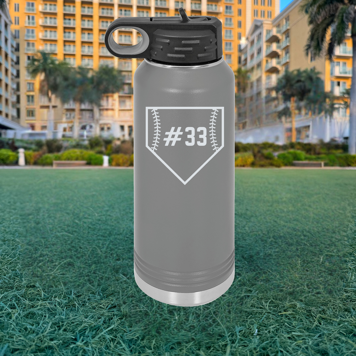 Gray stainless steel water bottle with black flip-top lid, engraved with sample baseball home plate design containing number 33, positioned on grass field with buildings in background.