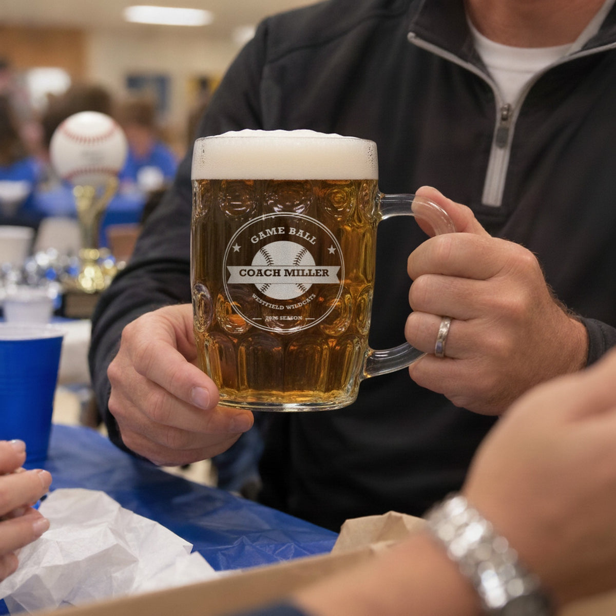 Baseball coach raising an engraved Game Ball beer stein at the end-of-season team banquet, a team trophy on the table behind him