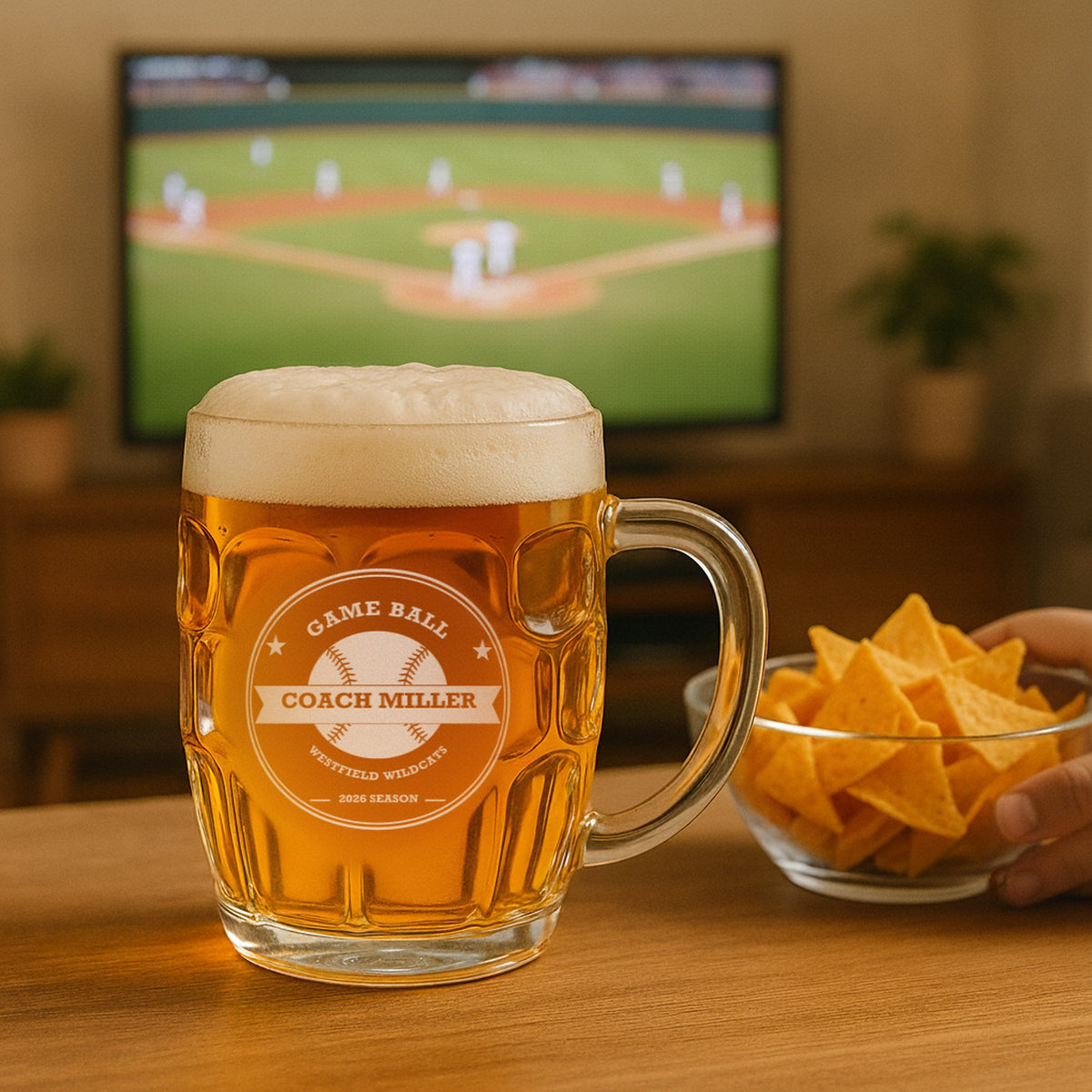 Personalized baseball coach beer stein filled with lager on a coffee table with chips, a ballgame playing on the television in the background