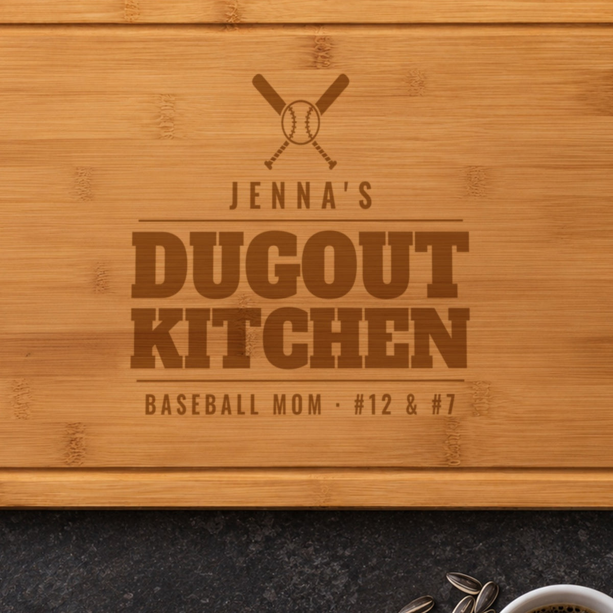 Close-up of the laser engraved baseball mom cutting board: crossed bats, a baseball, JENNA'S and DUGOUT KITCHEN burned into the bamboo