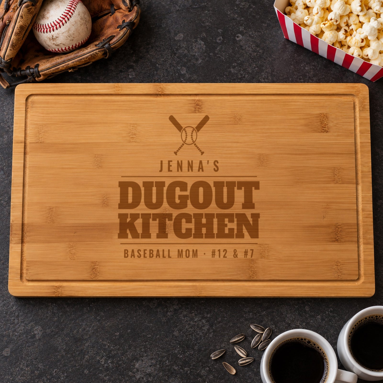 Baseball mom gift: bamboo cutting board engraved with crossed bats, JENNA'S DUGOUT KITCHEN and BASEBALL MOM #12 & #7, beside a baseball glove and popcorn