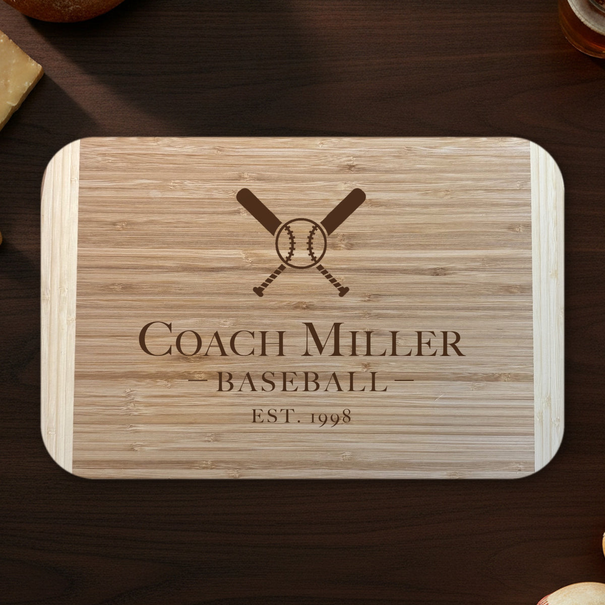 Baseball coach cutting board on a dark walnut counter with bread, cheese and a glass of beer, engraved Coach Miller BASEBALL EST. 1998 under crossed bats on two tone bamboo.