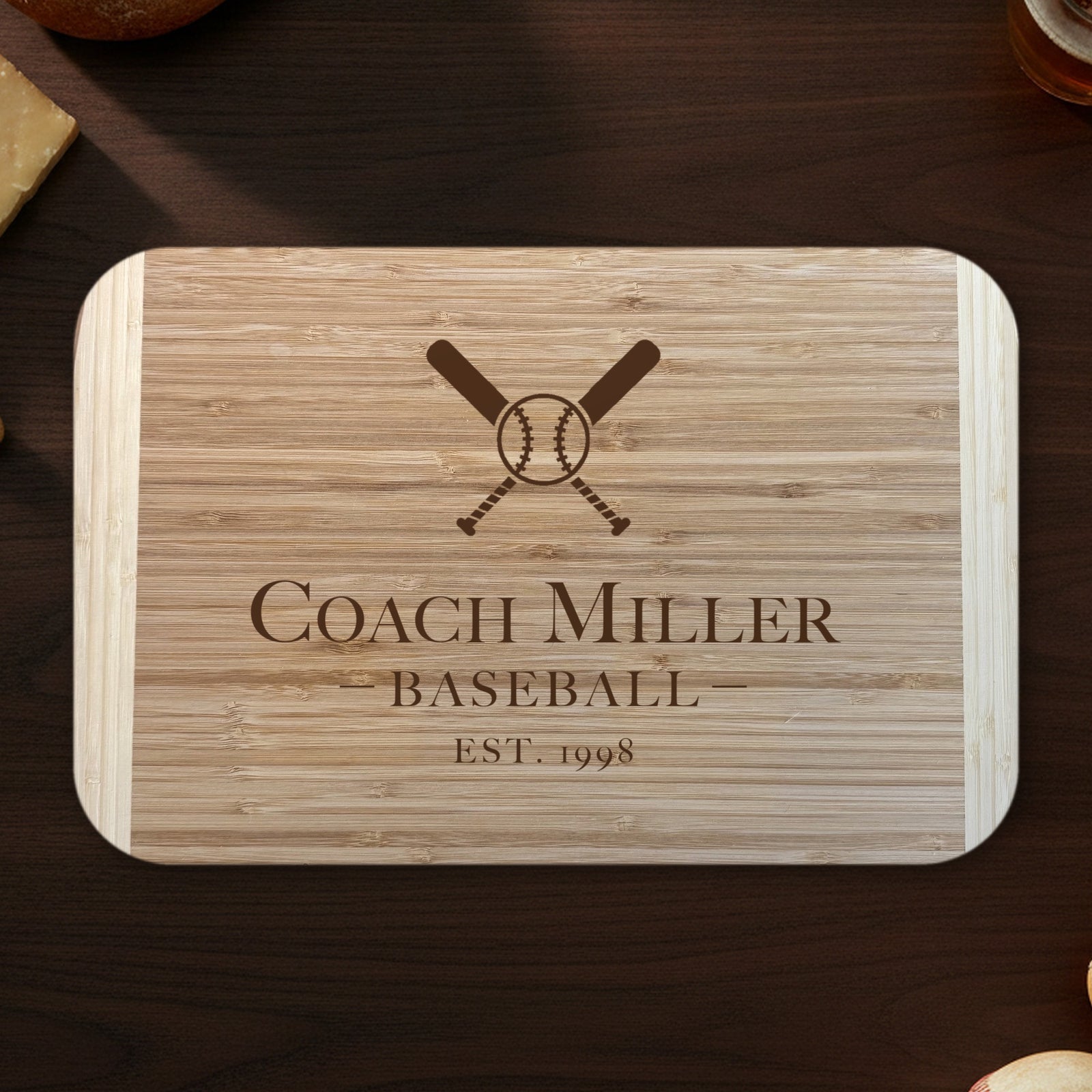 Baseball coach cutting board on a dark walnut counter with bread, cheese and a glass of beer, engraved Coach Miller BASEBALL EST. 1998 under crossed bats on two tone bamboo.