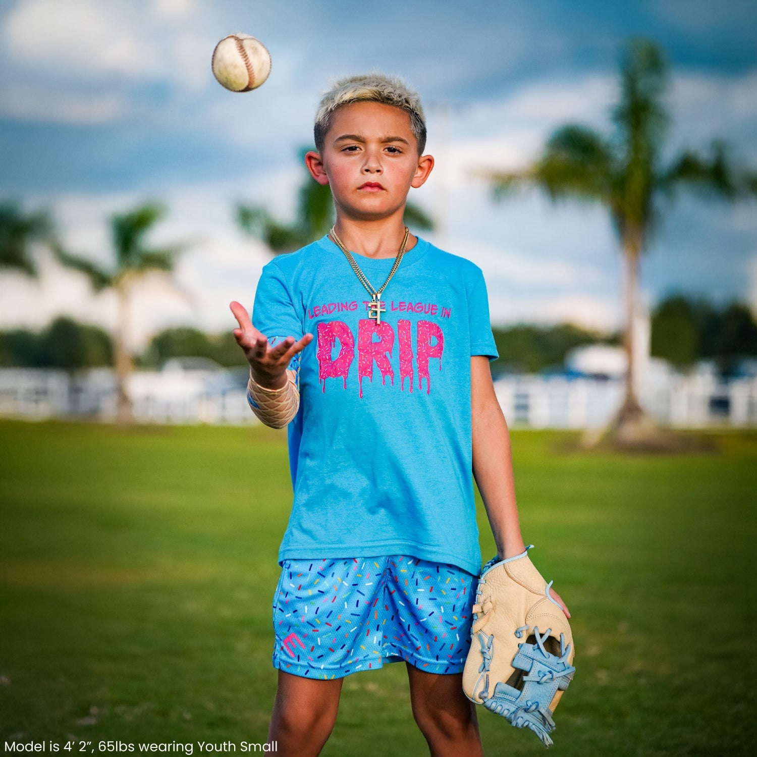 Child wearing turquoise Blue Sprinkles shorts with matching baseball-themed t-shirt, holding baseball glove, with baseball in air above head on grass field with palm trees and waterfront in background.