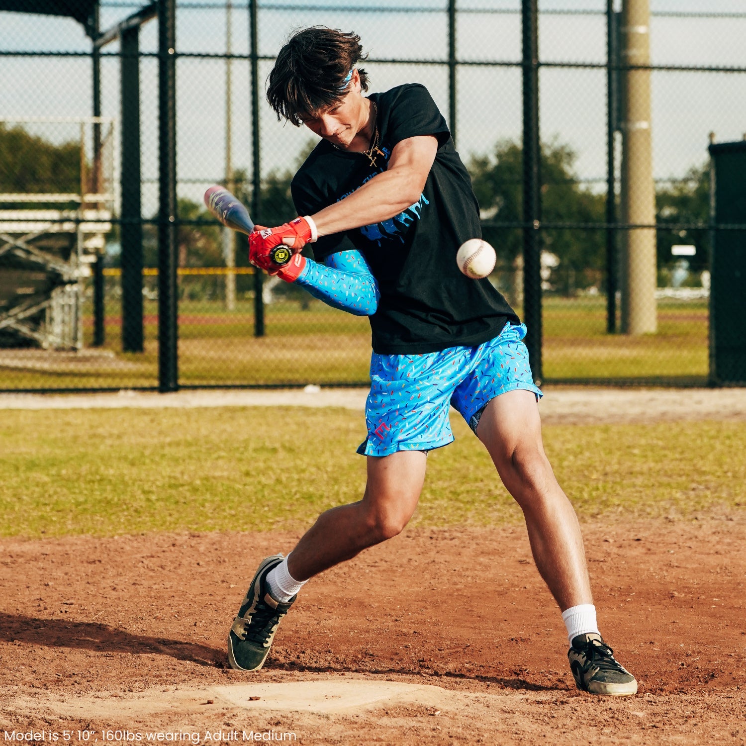 Person batting on baseball field wearing blue patterned shorts with black shirt and red batting gloves