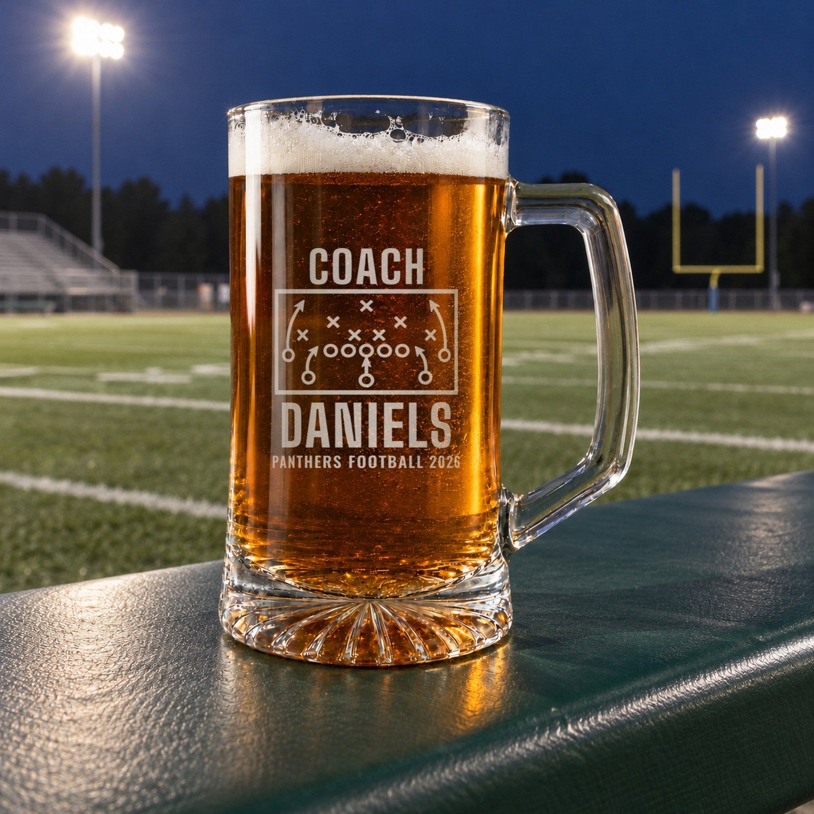 Engraved football coach beer stein full of lager on a stadium rail under Friday night lights, etched COACH, a play diagram, DANIELS and PANTHERS FOOTBALL 2026