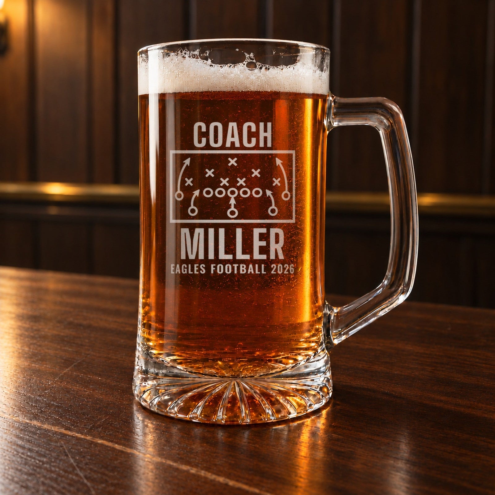Personalized football coach beer stein on a dark walnut bar, engraved with a play diagram, COACH, MILLER and EAGLES FOOTBALL 2026 in frosted glass