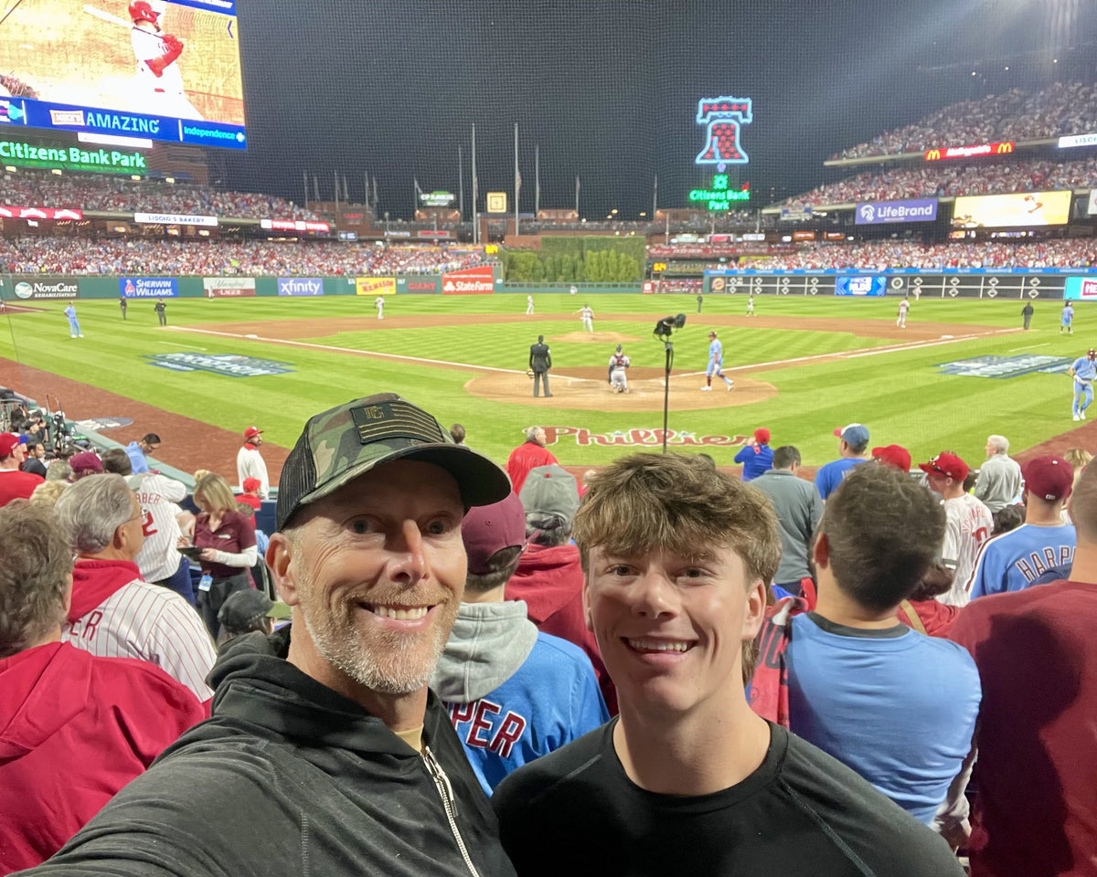 Chris Bajda and his son Tobey smiling in the stands at a night baseball game at Citizens Bank Park, the father and son behind Batter Box Sports