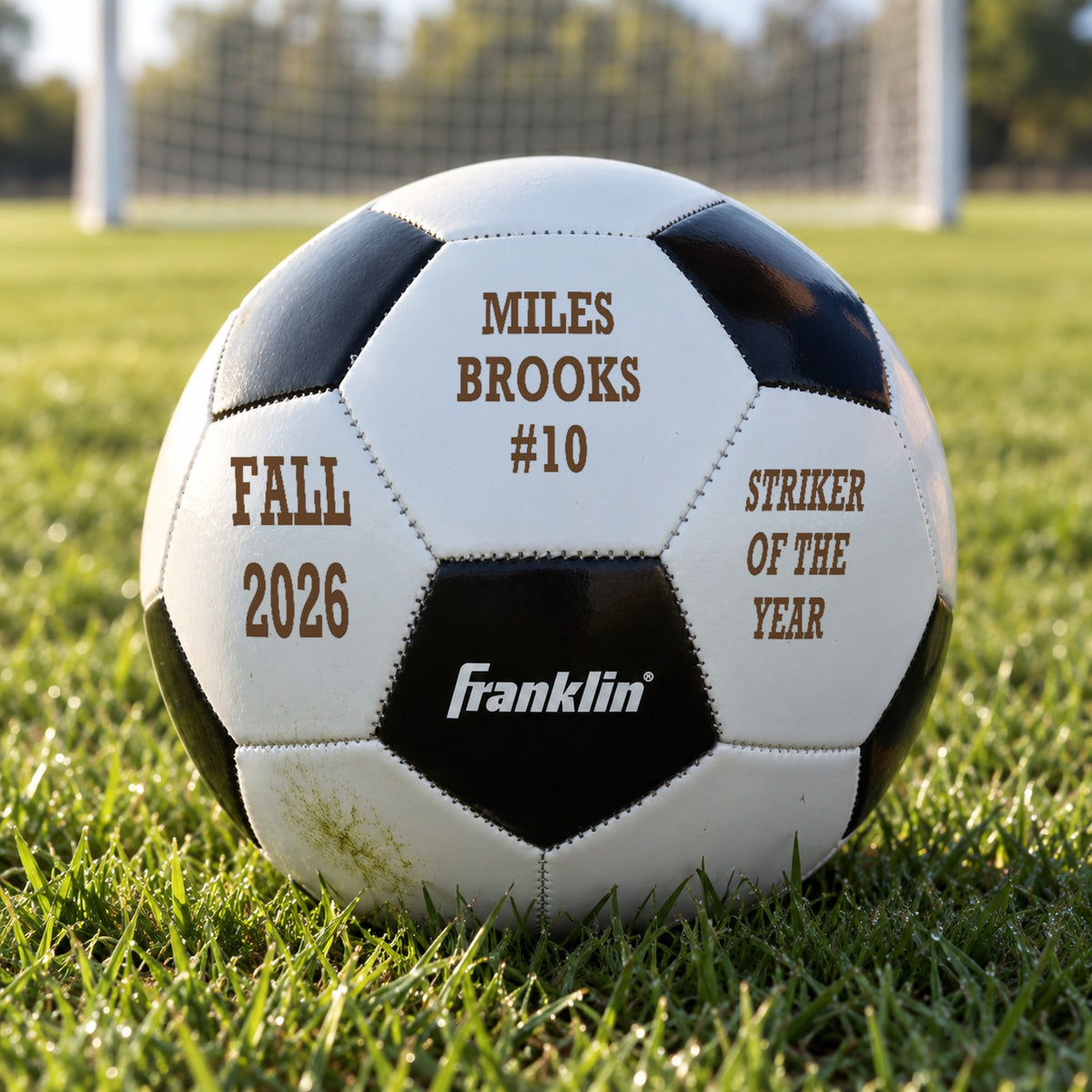 Custom soccer ball engraved with a player's name and #10, Fall 2026 and Striker of the Year, resting in the grass in front of a goal