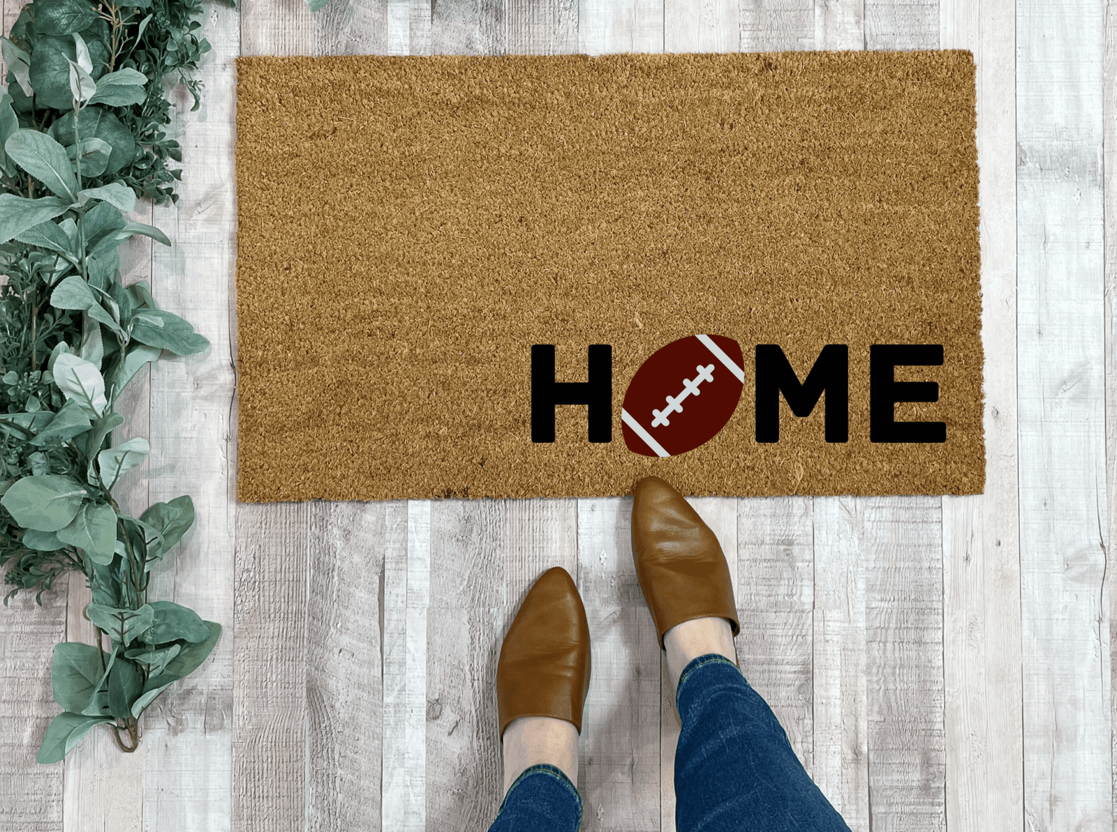 Tan cork doormat with black "HOME" text and brown football graphic replacing the O, displayed on whitewashed wood flooring next to potted greenery
