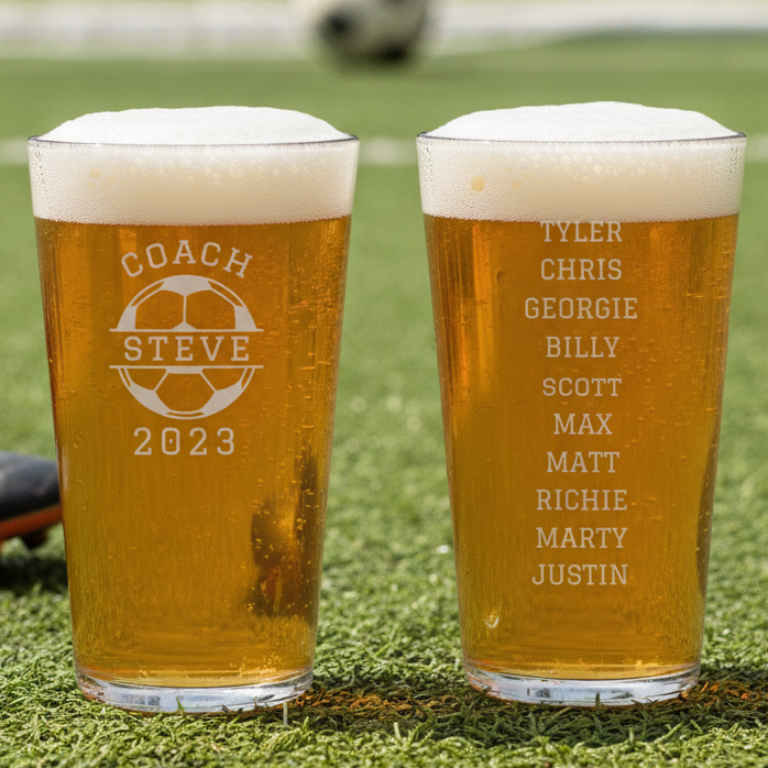 Two engraved pint glasses with beer, one showing sample coach name and soccer ball design, the other listing sample team member names, set on grass field