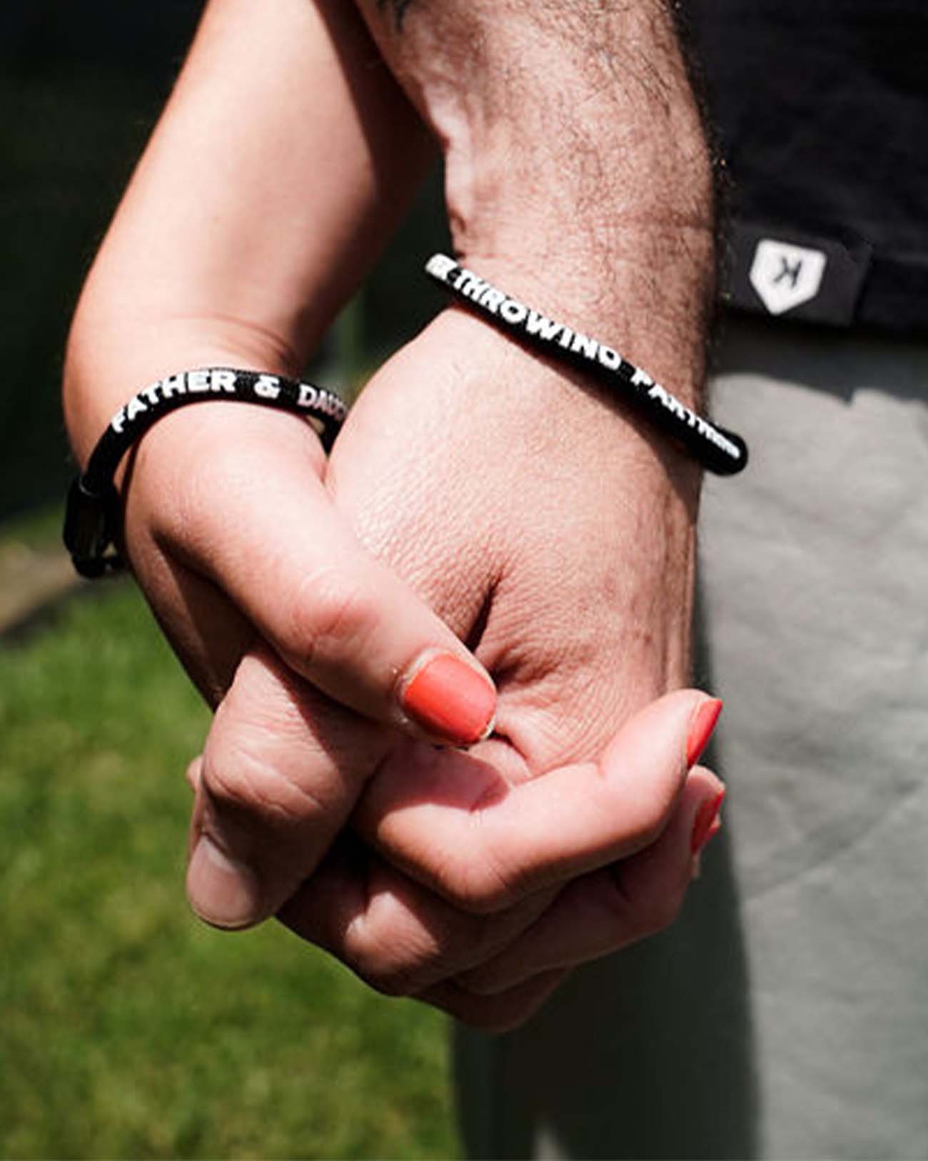 Two hands holding together, each wearing a black nylon bracelet with white screen-printed text reading sample names and "Forever Throwing Partners