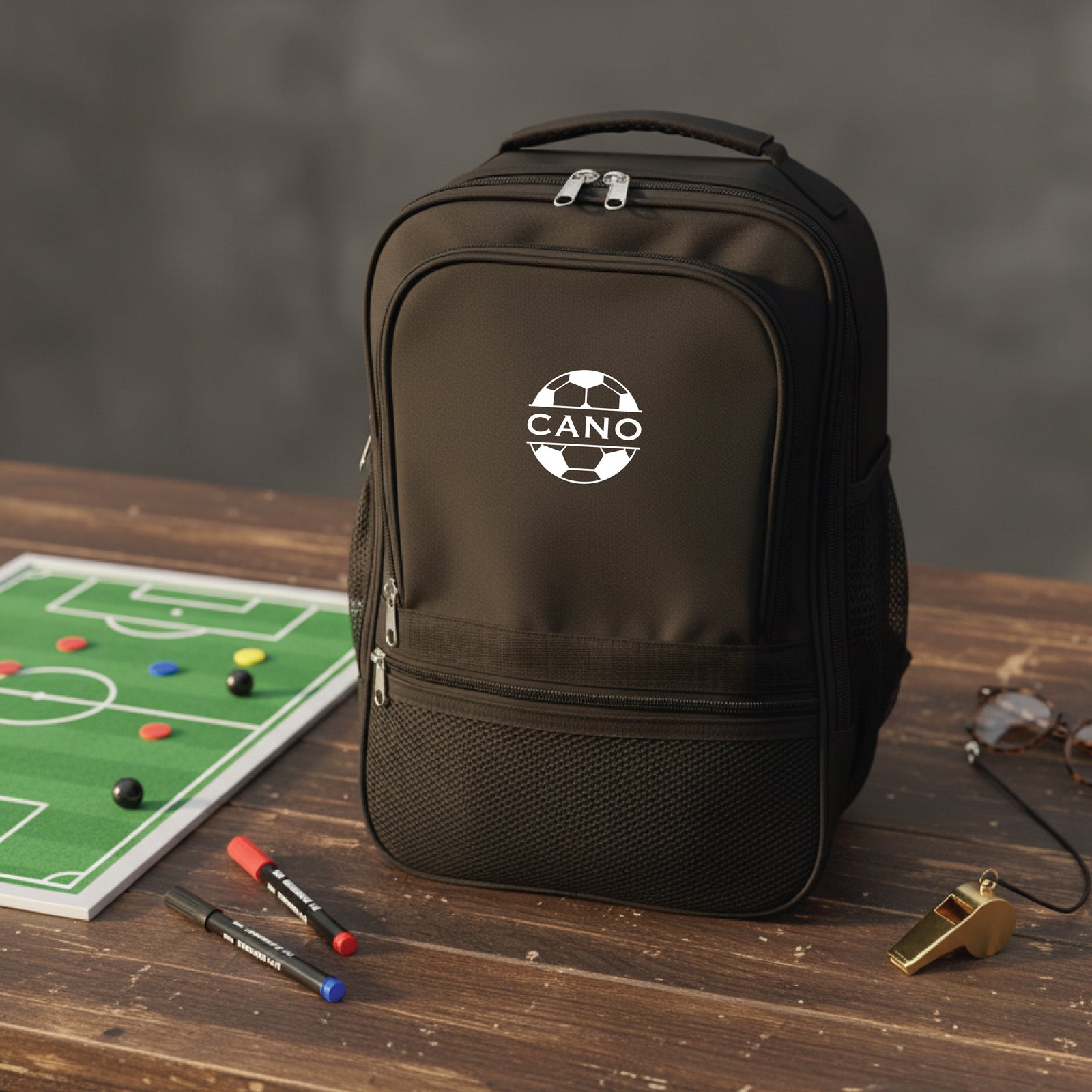 Black backpack with soccer ball logo and "CANO" text, displayed on wooden surface next to soccer field diagram, markers, and brass whistle