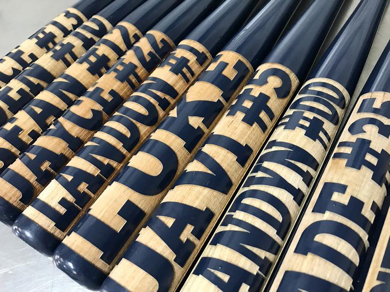 Navy mini baseball bats engraved with sample player names and jersey numbers, the two-line engraving layout used for first home run details