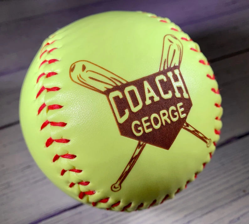 Neon yellow regulation softball with red stitching and laser-engraved sample design reading "COACH GEORGE" with a bat graphic in brown ink.