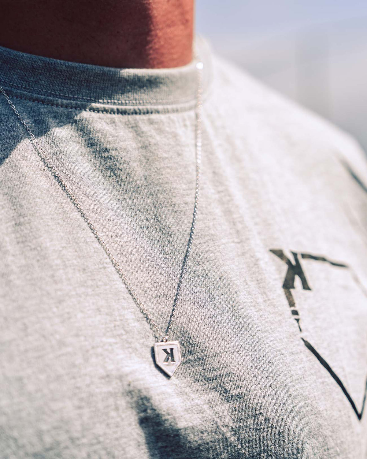 Silver K pendant necklace worn on chest, showing backwards K design on shield-shaped pendant with cable chain, styled on light gray t-shirt