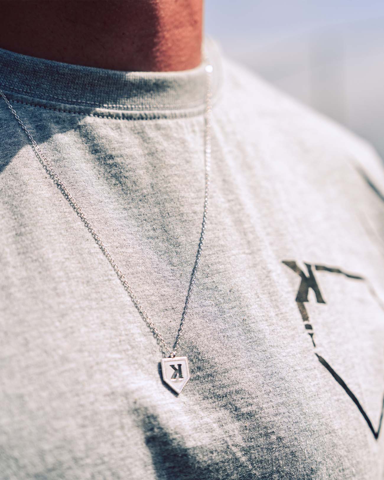Silver K pendant necklace worn on chest, showing backwards K design on shield-shaped pendant with cable chain, styled on light gray t-shirt