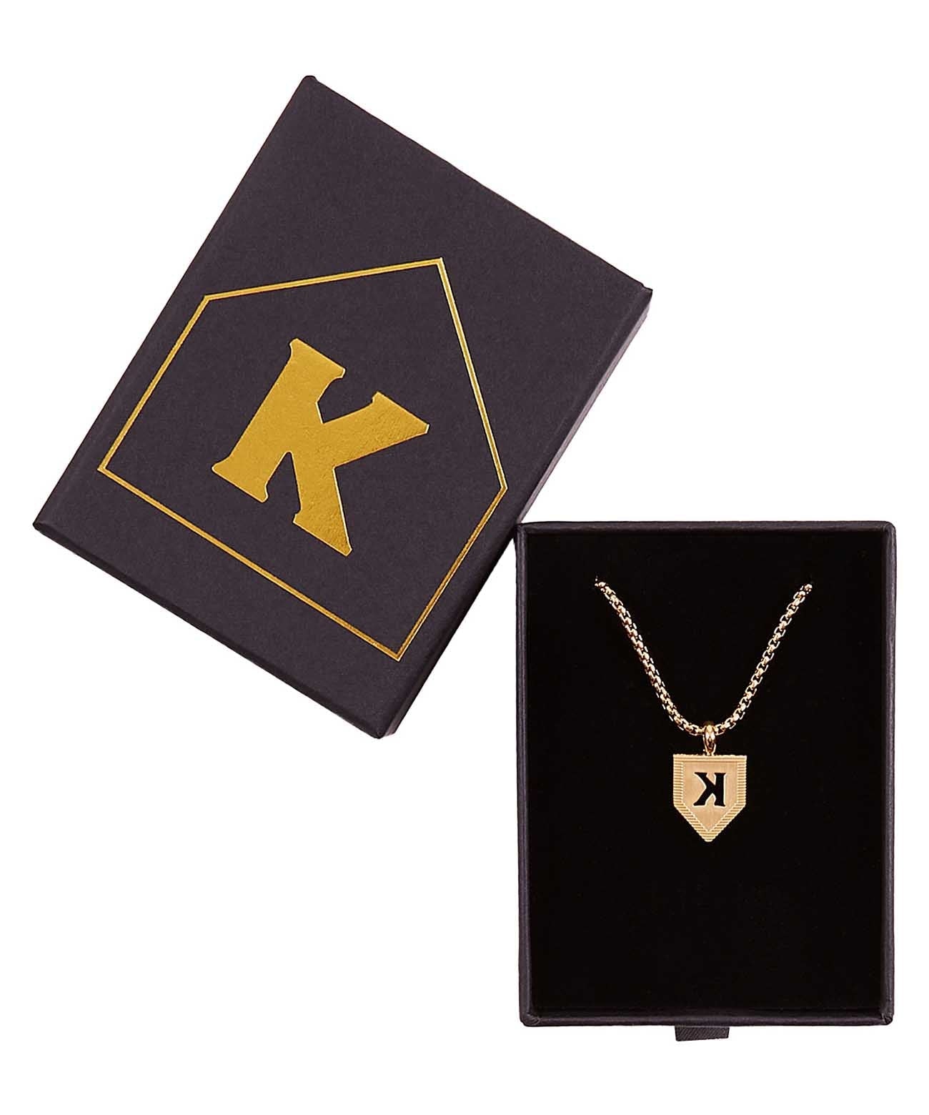 Dark navy jewelry box with gold K logo on lid, opened to reveal gold-plated pendant necklace with backwards K design on chain inside black-lined compartment.
