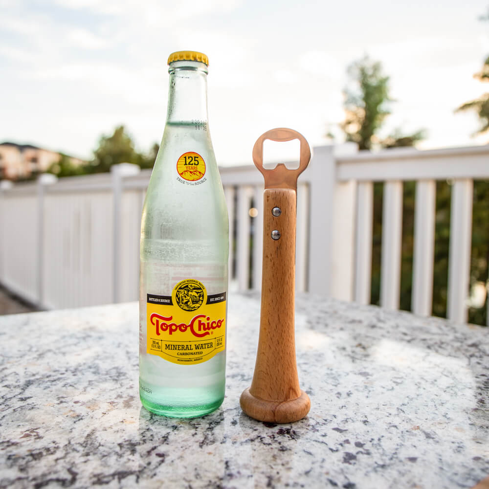 Wood baseball-bat handle bottle opener standing beside a Topo Chico bottle on a granite patio table outdoors
