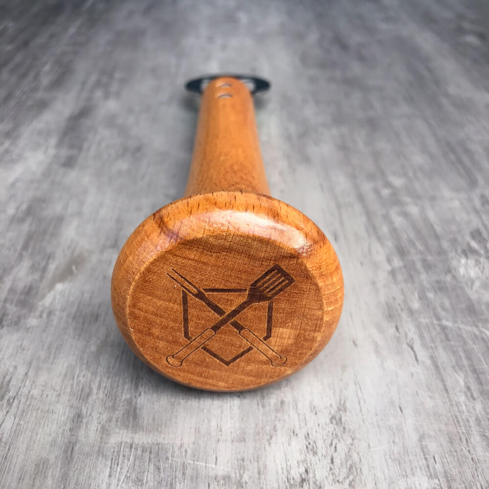 Close-up of the bat-knob end engraved with a sample design of crossed BBQ tools inside a diamond on the maple handle