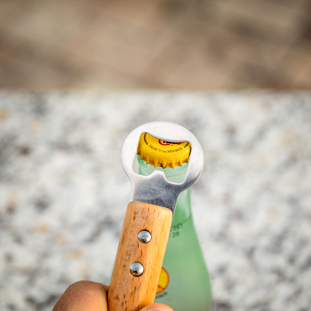 Hand using the baseball-bat bottle opener to pop a yellow cap off a glass soda bottle on a sunlit patio