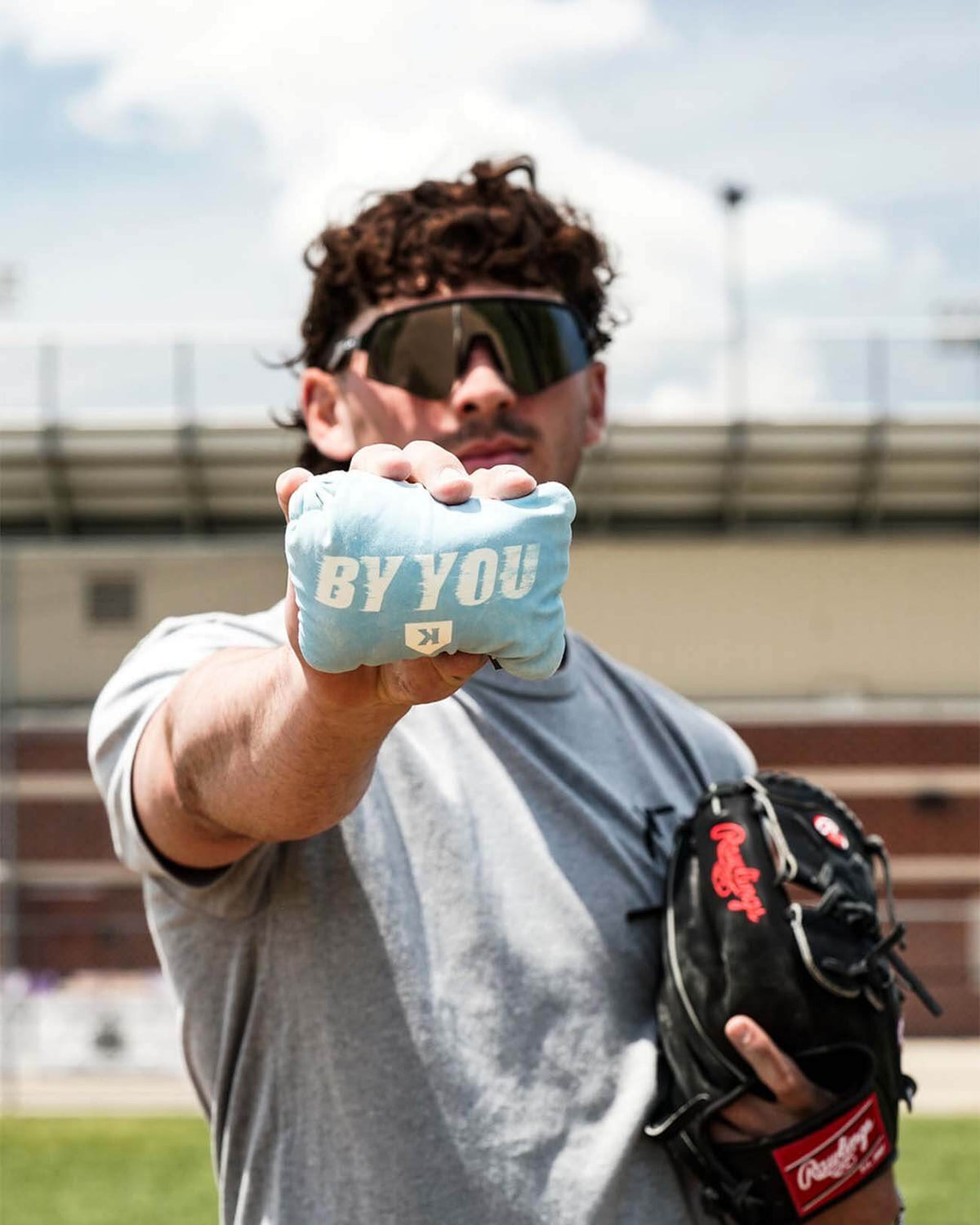 Person holding light blue rosin bag printed with sample text "BY YOU" at a baseball field, wearing dark sunglasses and baseball glove.