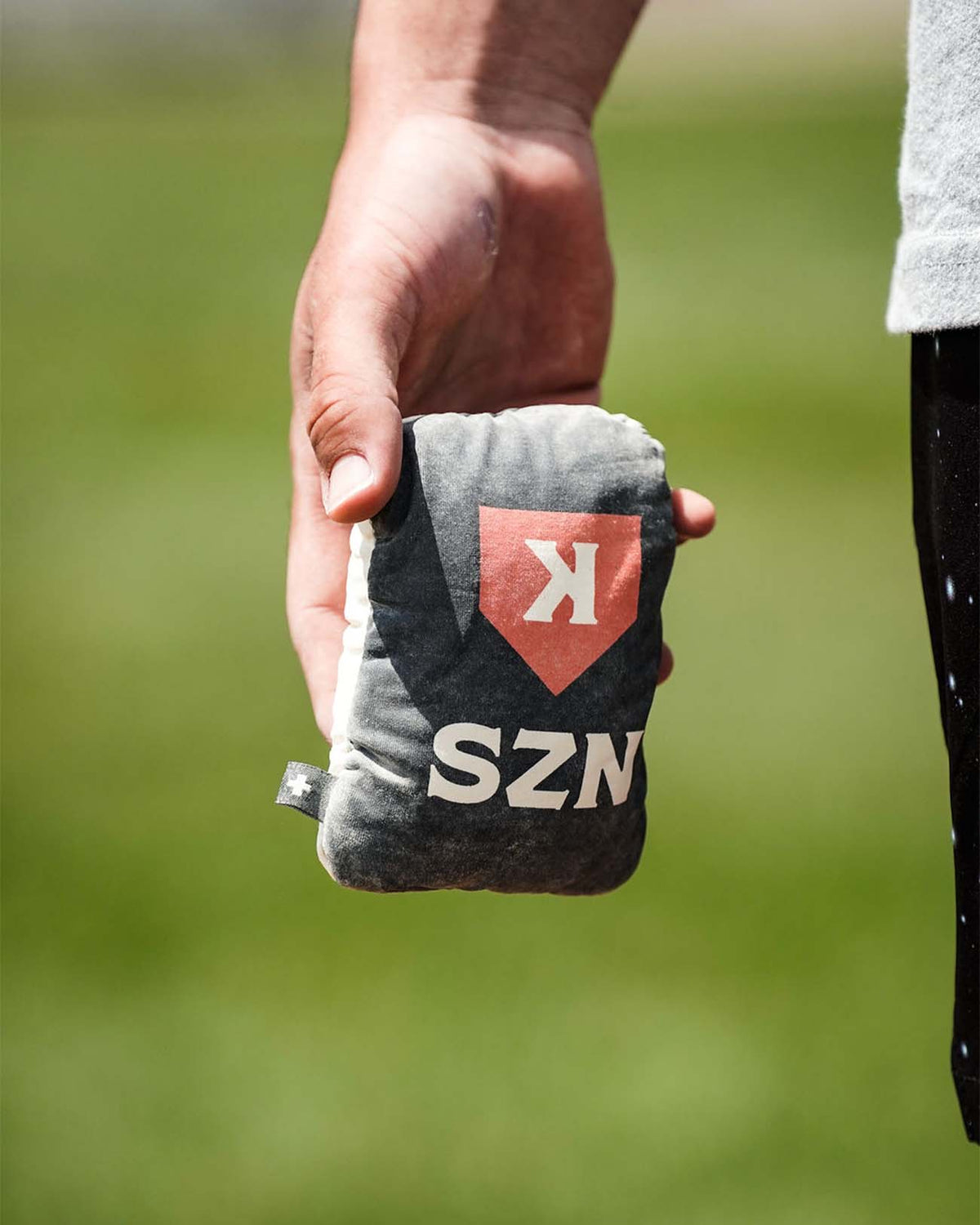 Hand holding gray rosin bag with red shield logo and white "K" and "SZN" text, displayed against blurred green field background