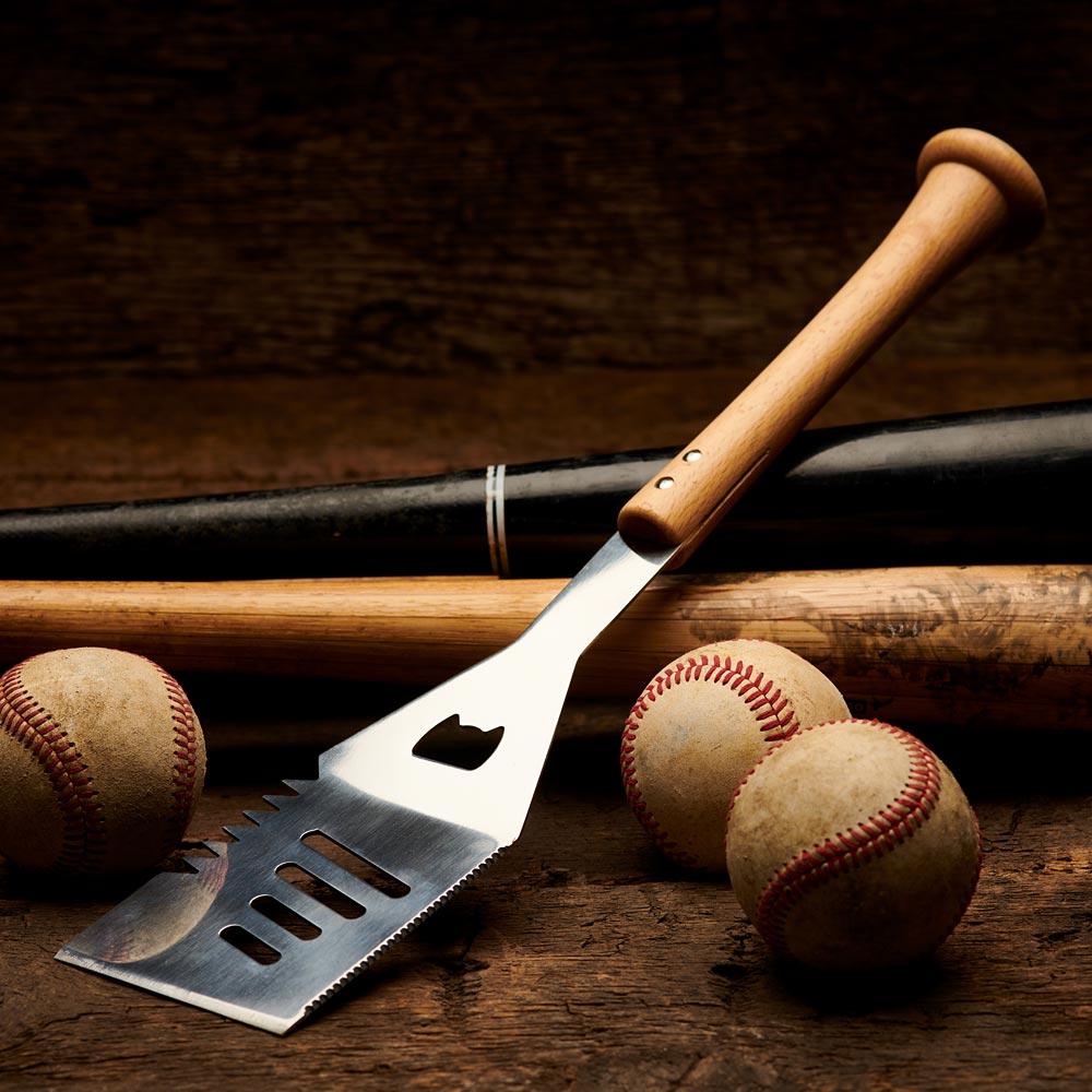 Five engraved bat-handle grill tools fanned across weathered wood planks beside a vintage glove and baseball, with sample player-name personalizations visible on each handle