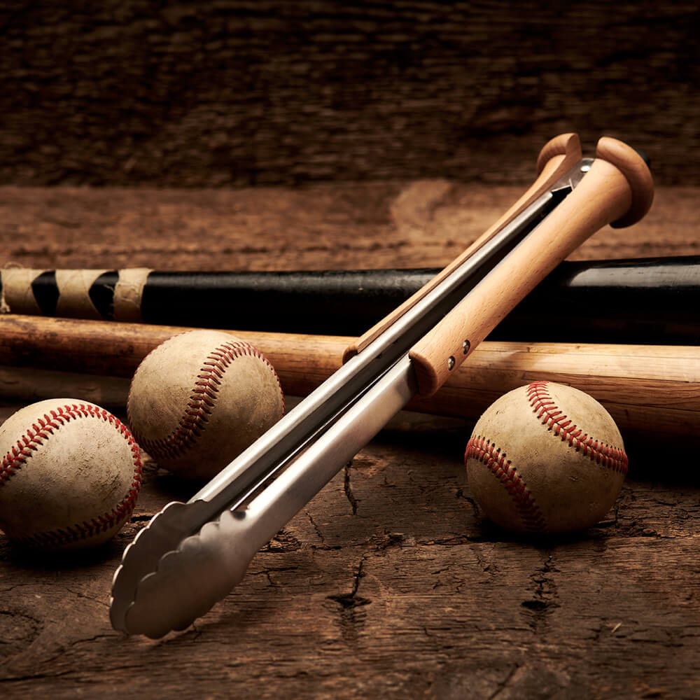 Close-up of four bat-handle grill tools engraved with sample 'COACH ROB / JUST HIT THE BALL. / DAMNIT! / COACH DON' personalizations on a grey concrete surface