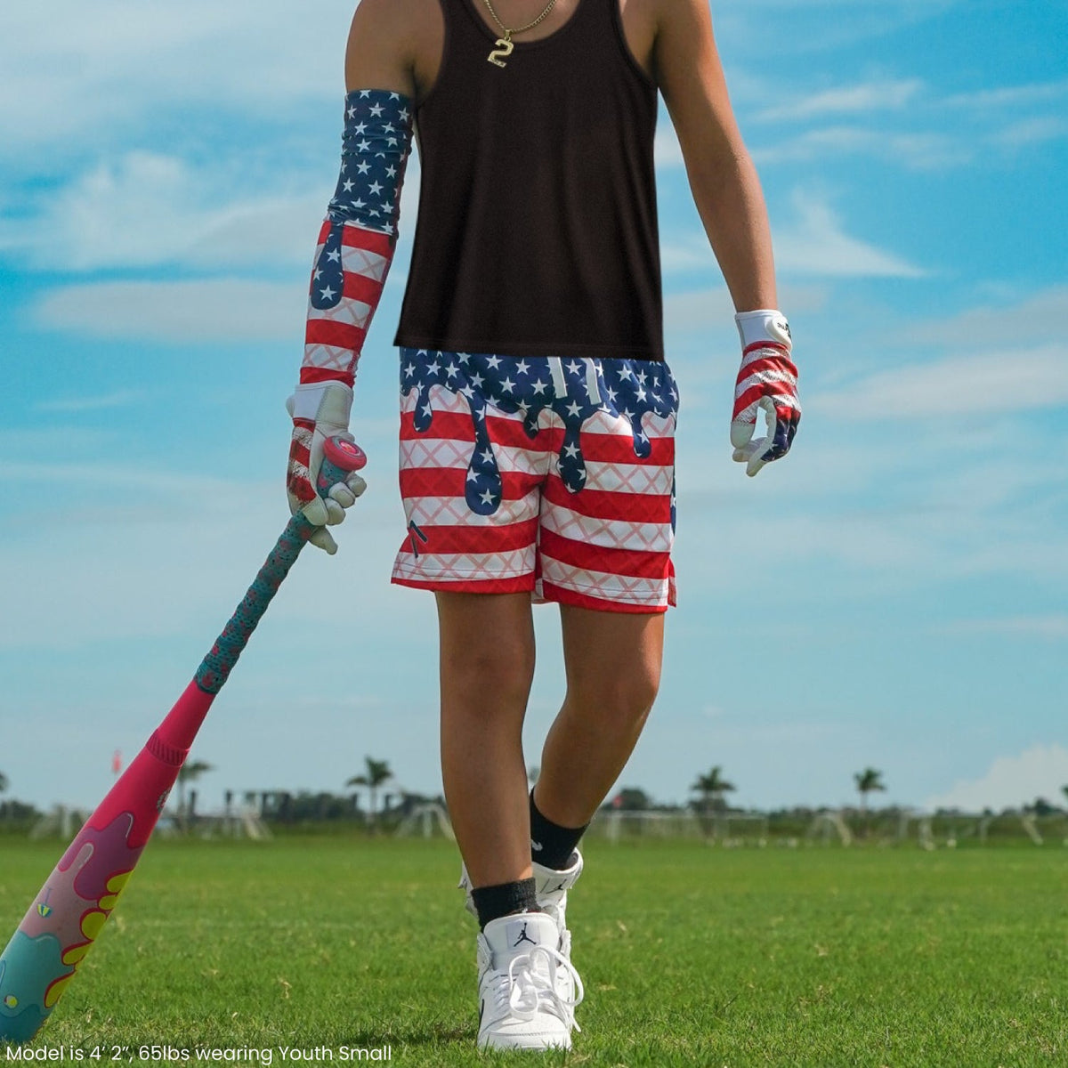 Athlete wearing USA flag patterned shorts with matching arm sleeve and glove, holding baseball bat on grass field under blue sky