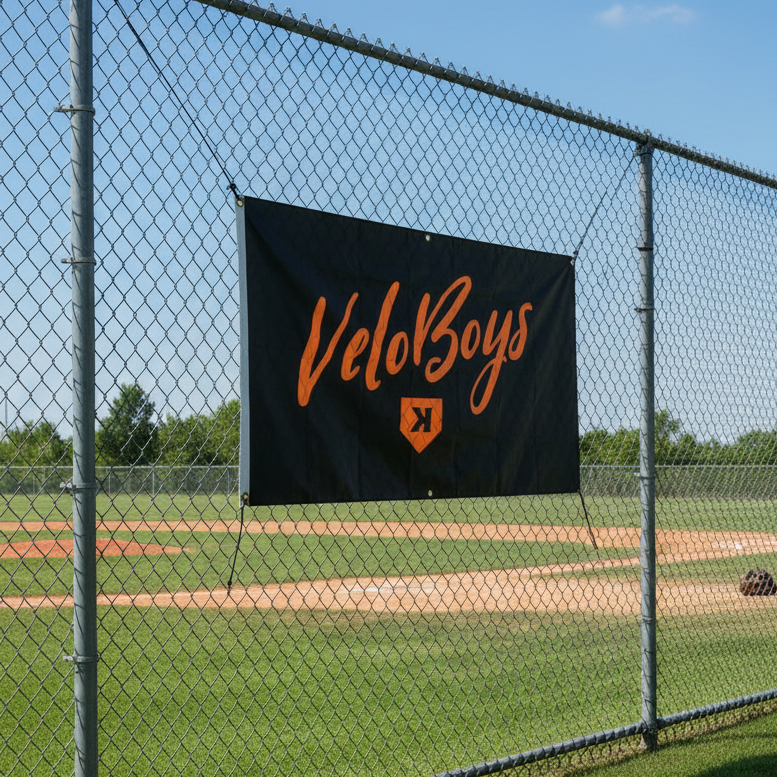 VeloBoys banner on a chain-link fence with a baseball field in the background