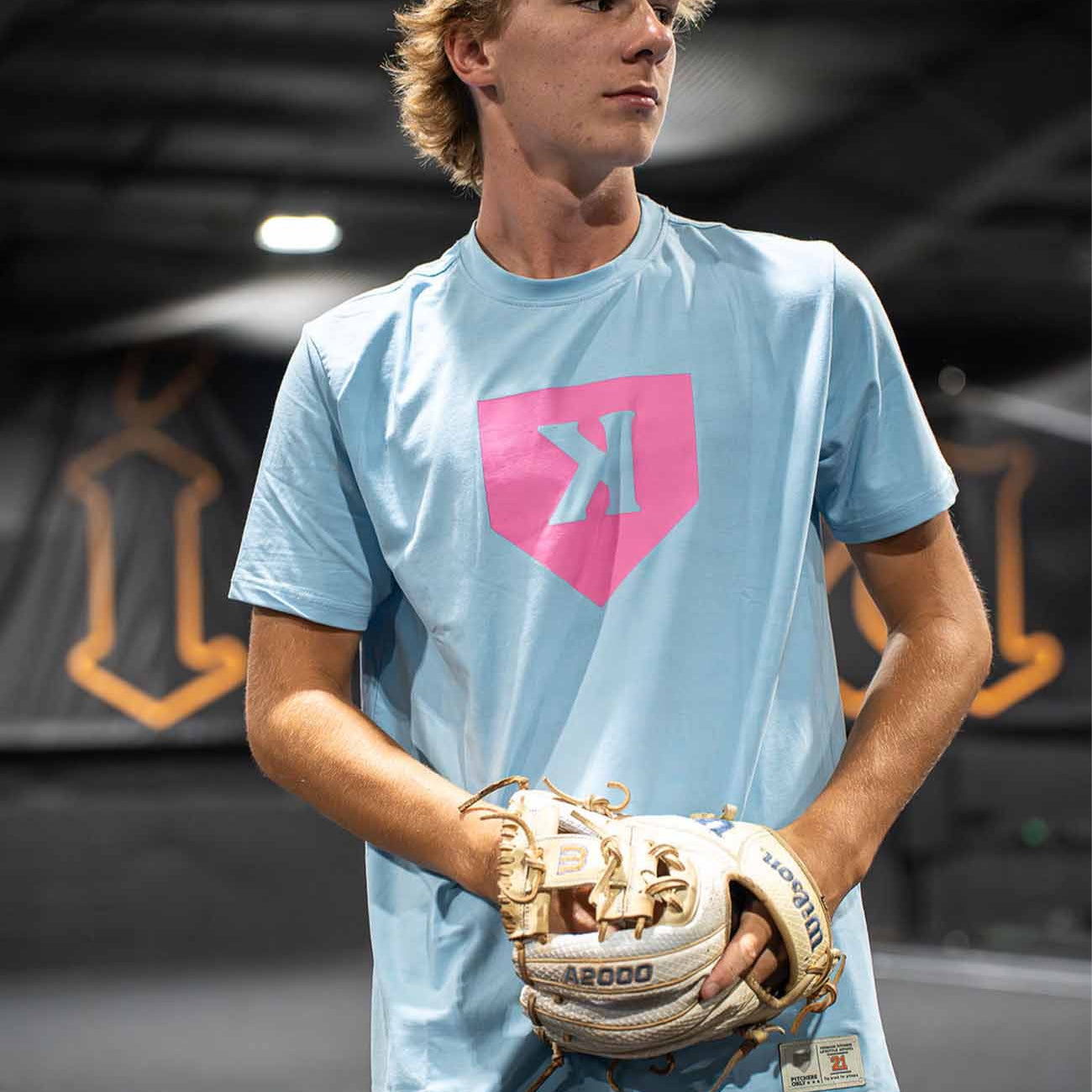 Person wearing a light blue t-shirt with a pink backward k on home plate, holding a baseball glove indoors.