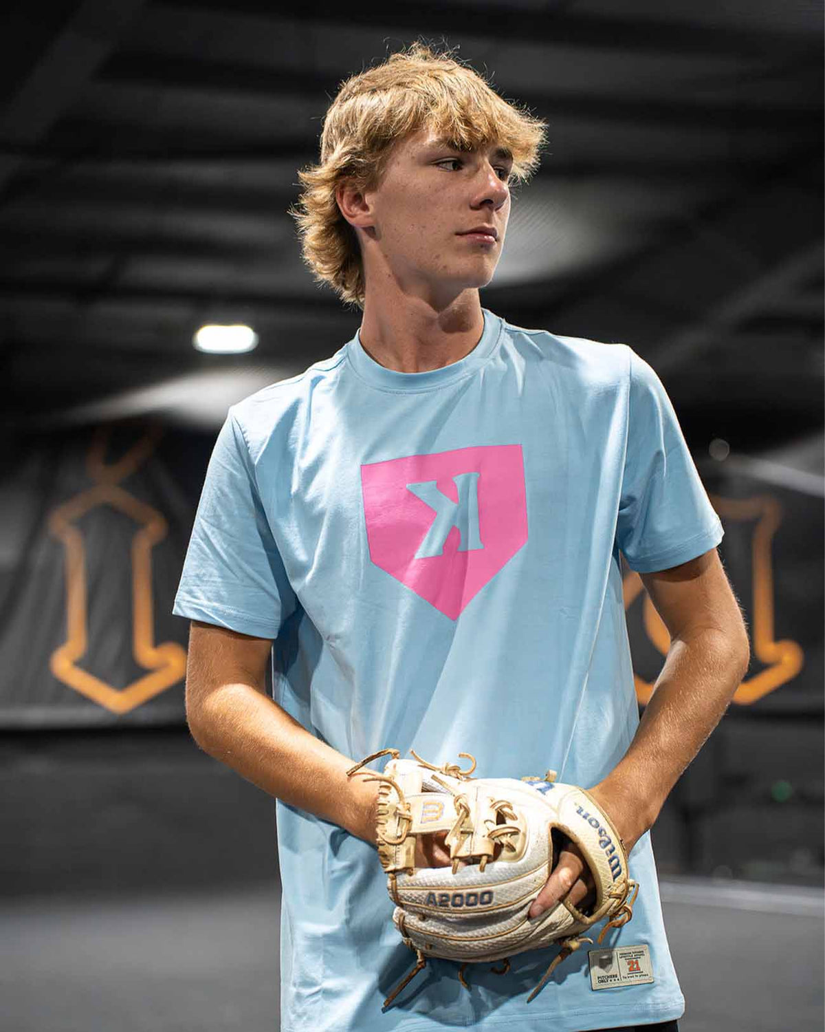 Young person wearing light blue fitted tee with pink home plate and stylized pitcher logo, holding tan and brown baseball glove in indoor facility