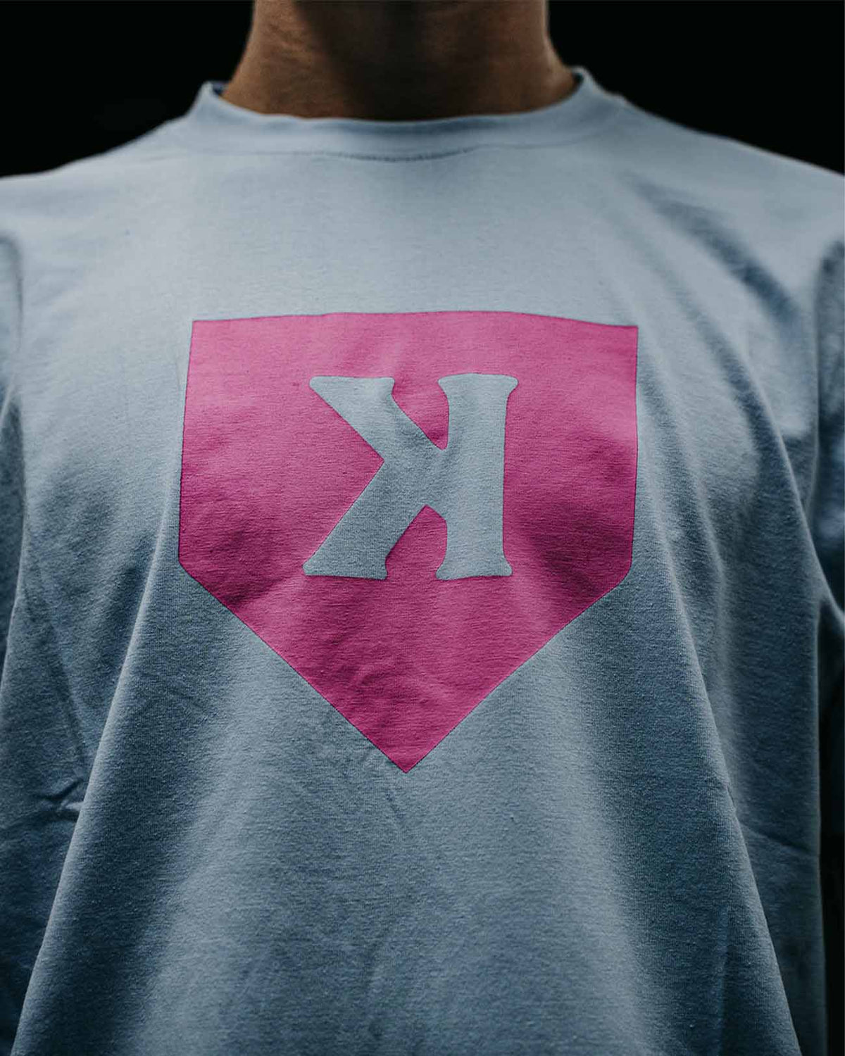 Light blue t-shirt with pink home plate graphic and gray letter X displayed on the front chest area