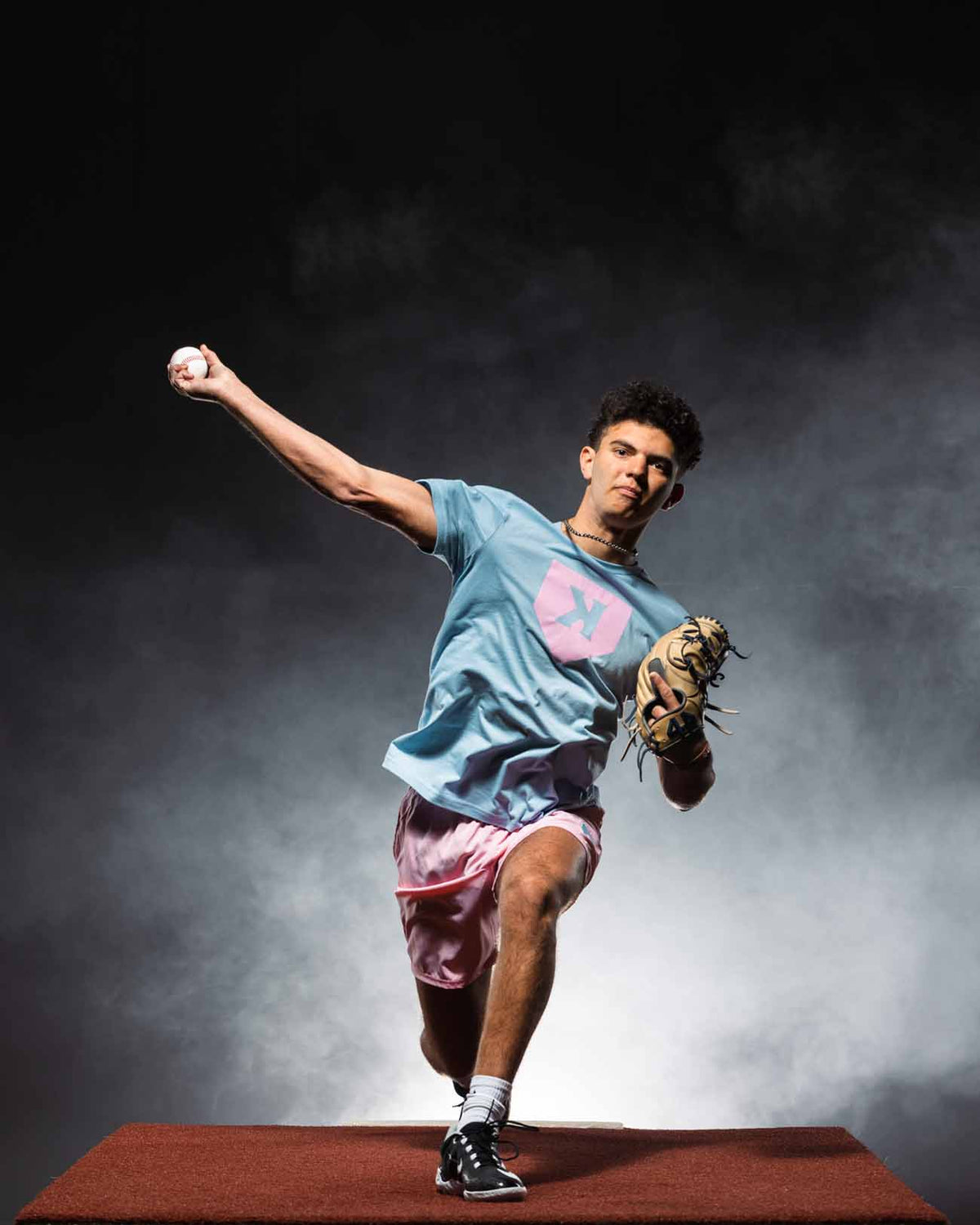 Young male pitcher in mid-delivery stance wearing cotton candy blue tee with pink logo and burgundy shorts, holding baseball and leather glove