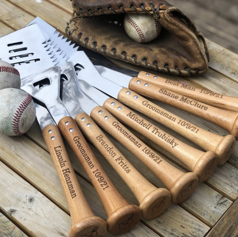 Five natural wood spatulas with laser-engraved sample names and dates arranged on whitewashed wood, displayed with baseballs, baseball glove, and grilling tools